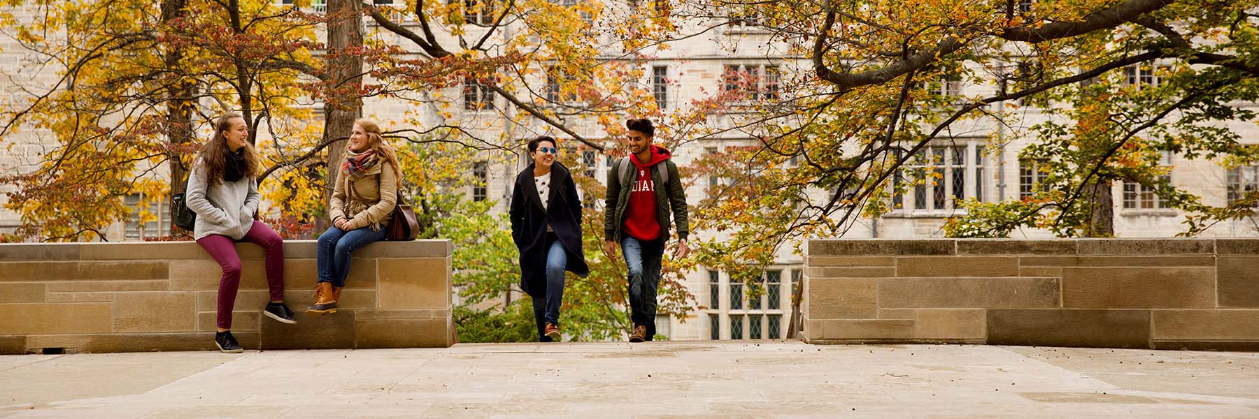 An autumn day in front of the Indiana Memorial Union, where two student sit laughing under the orange and yellow foliage of trees.