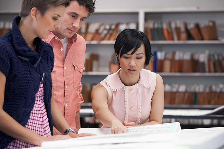 An open map is spread out on a desk; one student points at the map while two others look to see what she's pointing at.